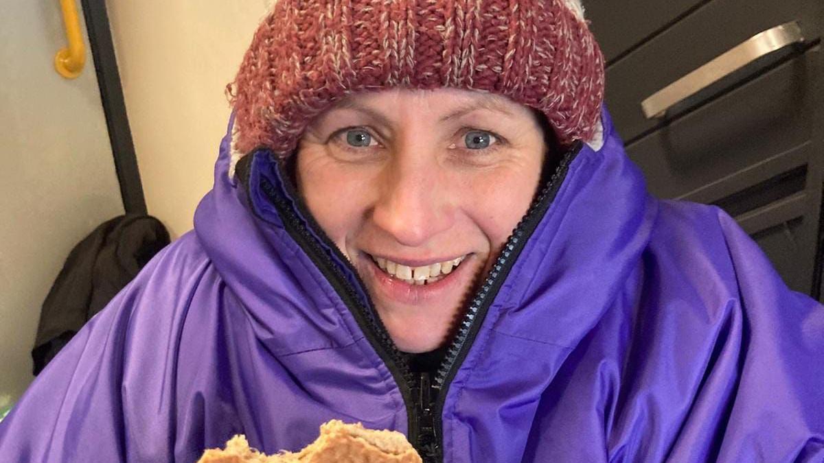 A woman wearing a red woolly hat and a bit purple coat, eating a waffle and smiling at the camera.