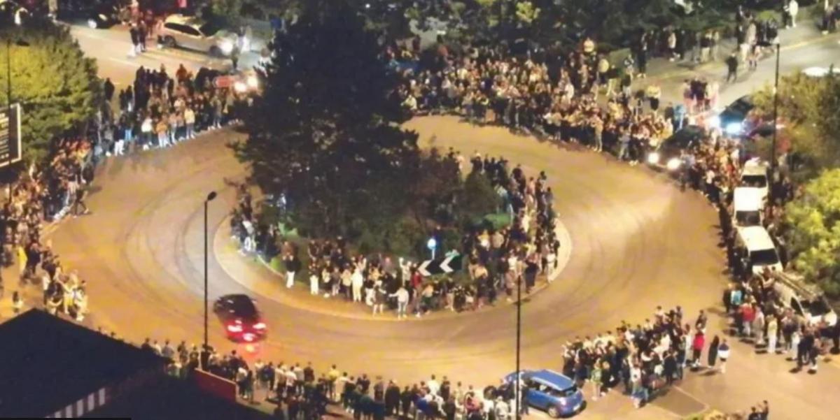 An overhead shot at night time of cars going around a roundabout whilst crowds line the pavement.