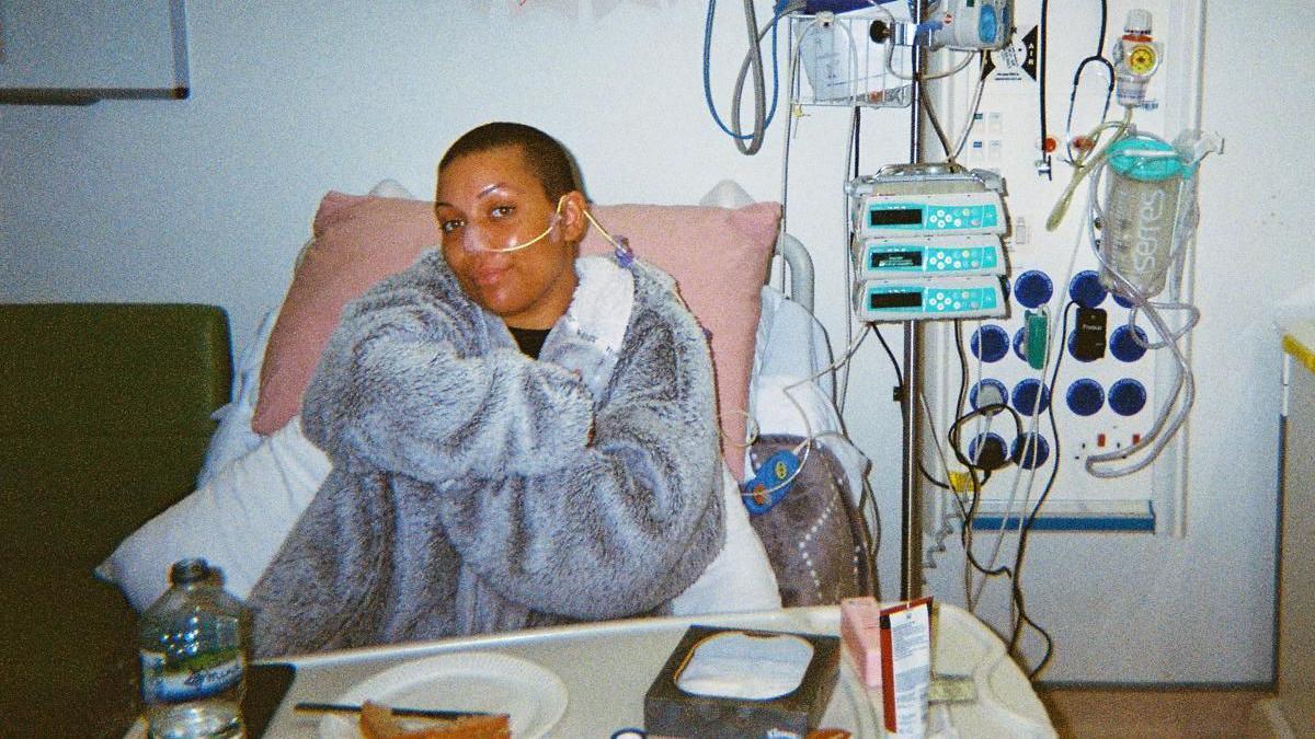 Sacha Gomez, in a hospital, undertaking treatment, wearing a grey fluffy top, sitting in a bed, with food in front of her and machinery to the right. She has a tube in her nose. She is smiling and looking at the camera. A white bin is to the right.