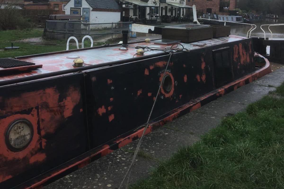 A narrowboat with circular windows painted black with patchy red paint. The craft sits next to a lock and alongside the concrete edge of a towpath. Behind it are several buildings.