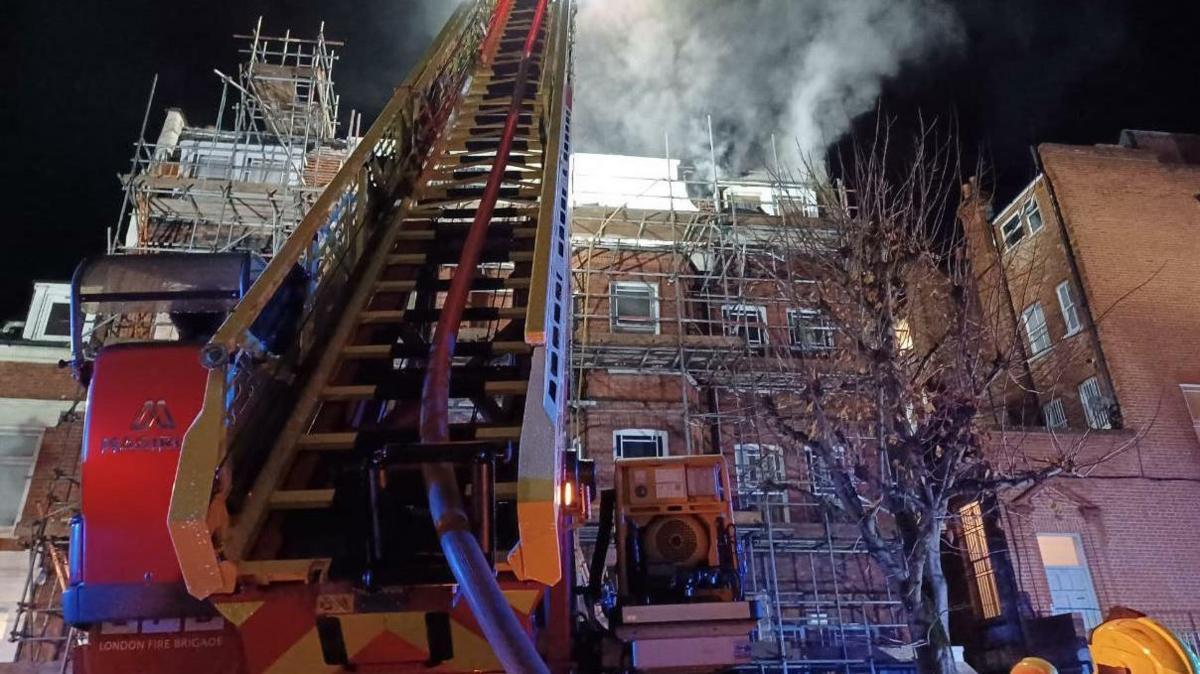 A red and silver turntable ladder stretches up and over a converted brick block of flats covered in scaffolding, with smoke rising into the dark sky