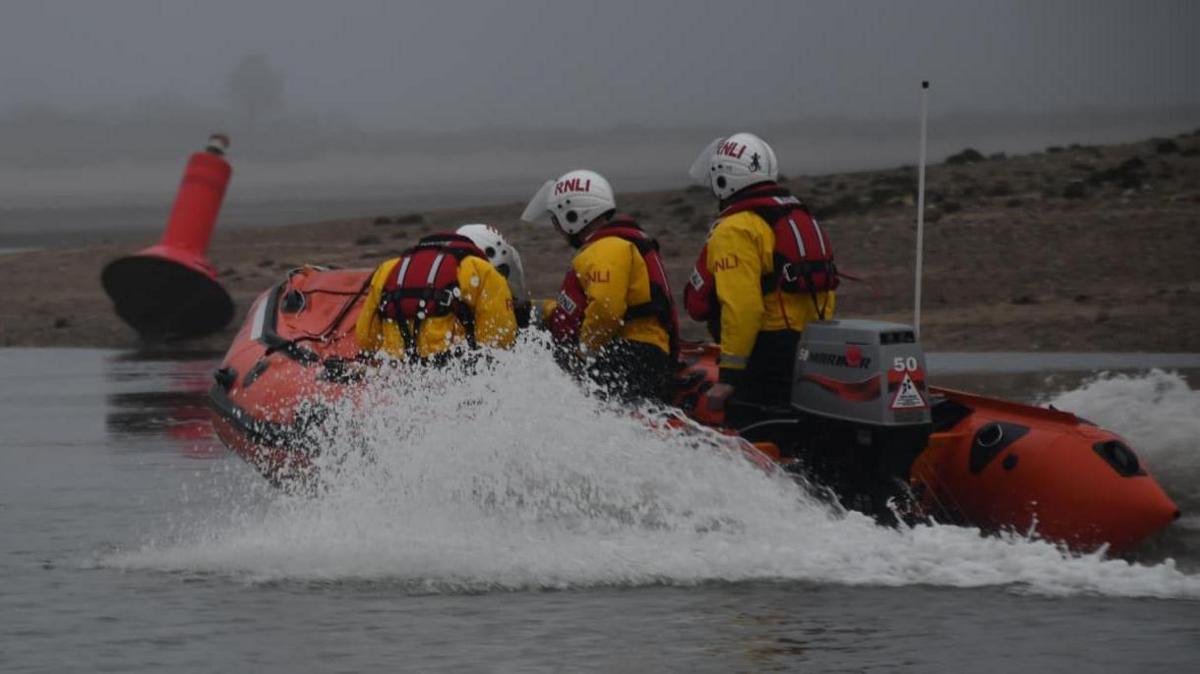 Three RNLI crew onboard an orange inshore lifeboat. They all wearing red life jackets, white helmets and yellow jackets. Water is spraying up the sides of the boat as it travels through the water in foggy conditions.