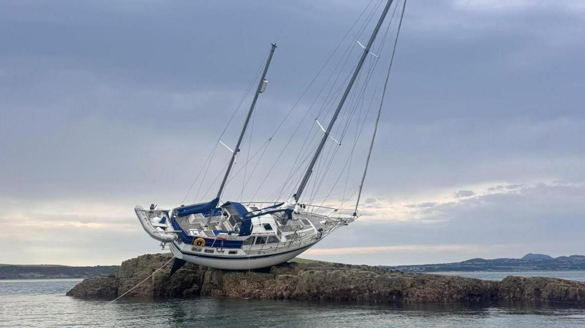 A wider shot of the yacht on its side on rocks in the sea, with cloudy skies behind
