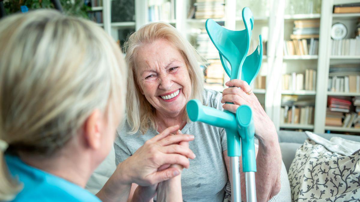 A healthcare worker with long blonde hair holding the hand of an older lady with crutches in her hair. She also has blonde hair and is wearing a grey t-shirt.