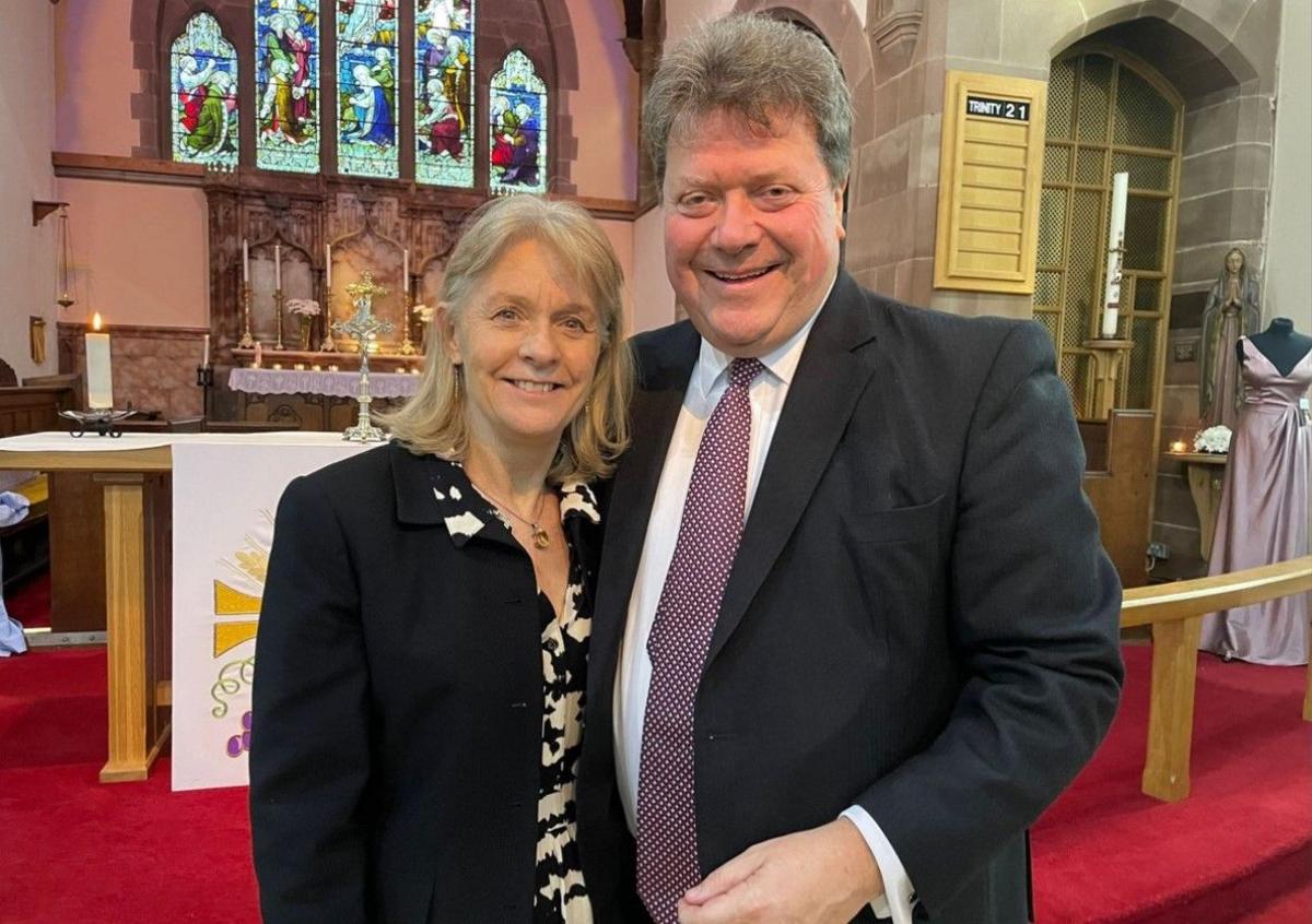 Sue and Alex Tatham inside the church with a stained glass window and candles behind them. Both are wearing jackets and standing on red carpet.