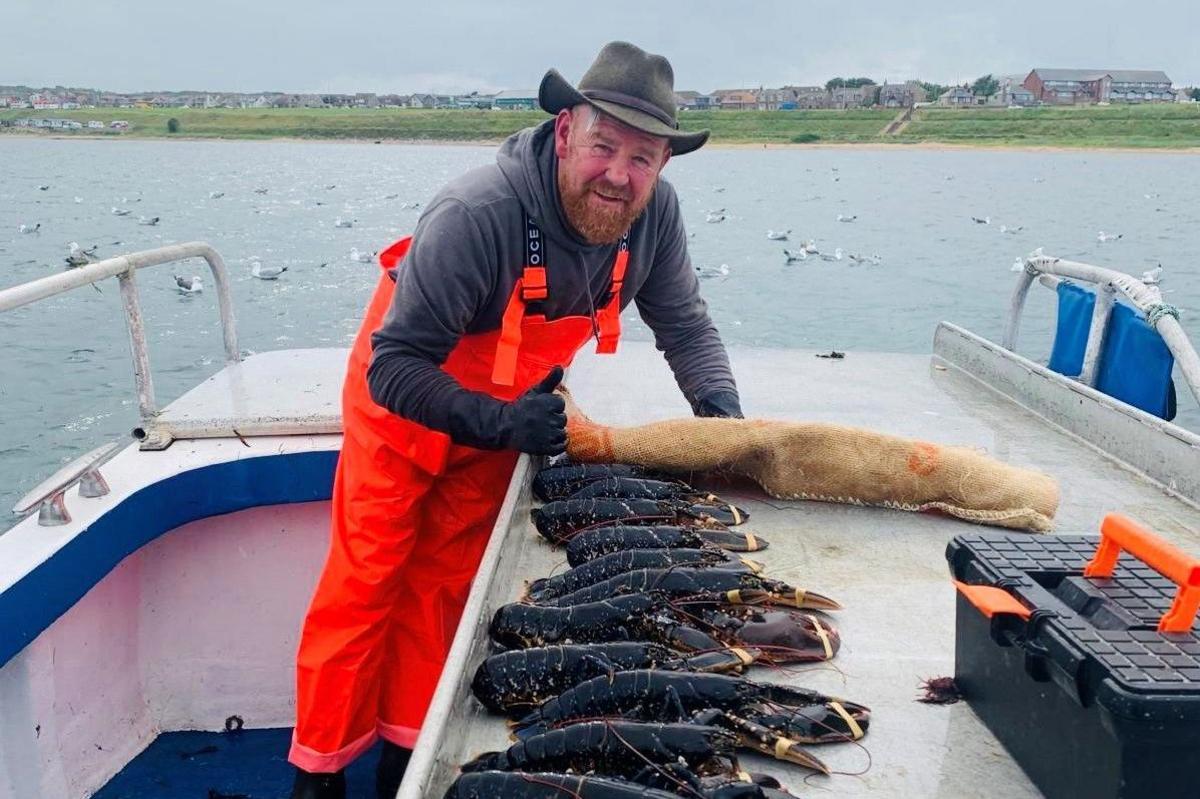 Man on boat wearing green cowboy-style hat, grey top and orange overalls, with several dark lobsters with their claws tied together.