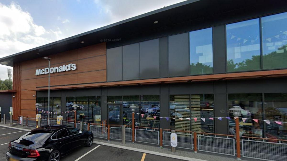 The outside of a McDonald's restaurant. It has dark wood and dark grey cladding and a fully glazed frontage. A car can be seen parked outside and bunting is hung between wooden lamp-posts.