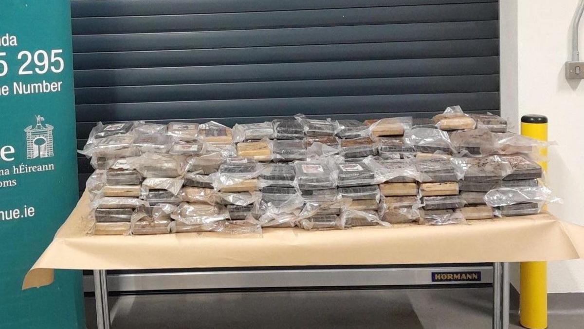 A close-up of a table placed in front of a grey shutter inside a non-descript room. The table has a light brown covering. On top are stacks of brown-looking material in plastic packaging, perhaps two rows with 12 stacks in each row. The drugs on the table are beside a partially-seen sign with the logo and contact details for the Irish Revenue service
