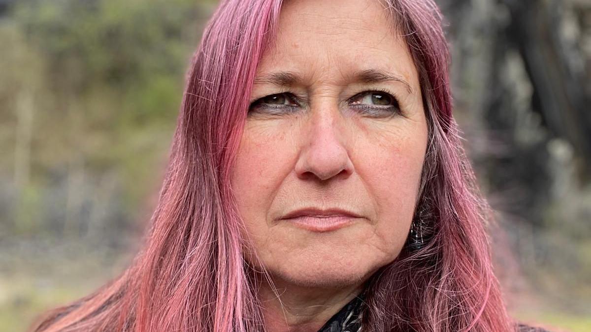 Helen looks into the distance, away from the camera, with a serious expression. She has long dyed pink hair, and is standing outside with blurred greenery behind her.