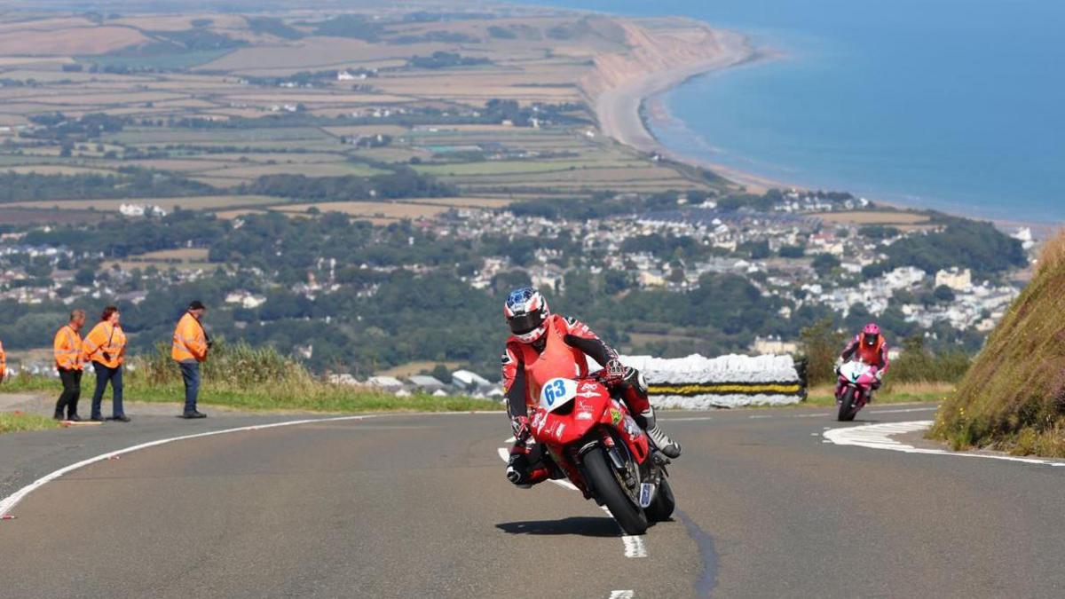 Two bikes on climb up the Mountain Road, one is red and is followed by another that is pink and there are marshals in orange jackets on the left. There is a picturesque scene behind them of green fields and the blue sea of the sweep of Ramsey Bay.