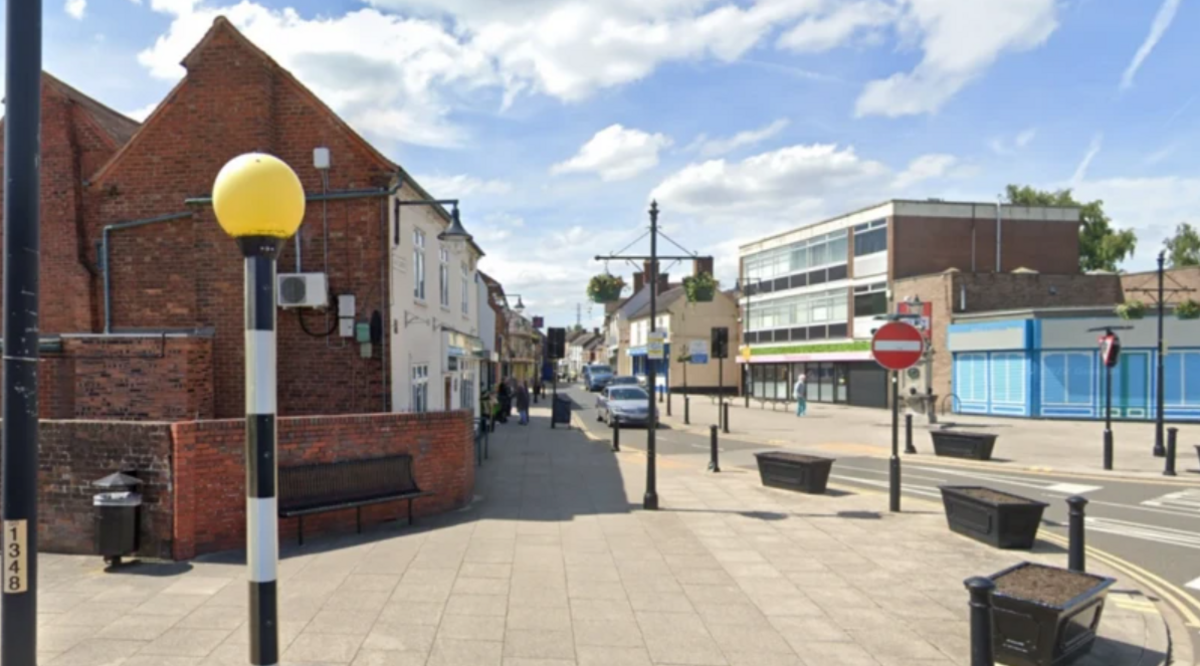 A view of a street with a pelican crossing light on the left hand side and a road running past a row of shops with a pub at the end