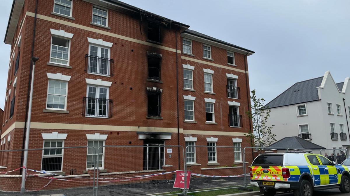 A picture of the outside of the flats. It's a large brick building, with fencing around it and a police vehicle in front it. There is signs and police tape. 
