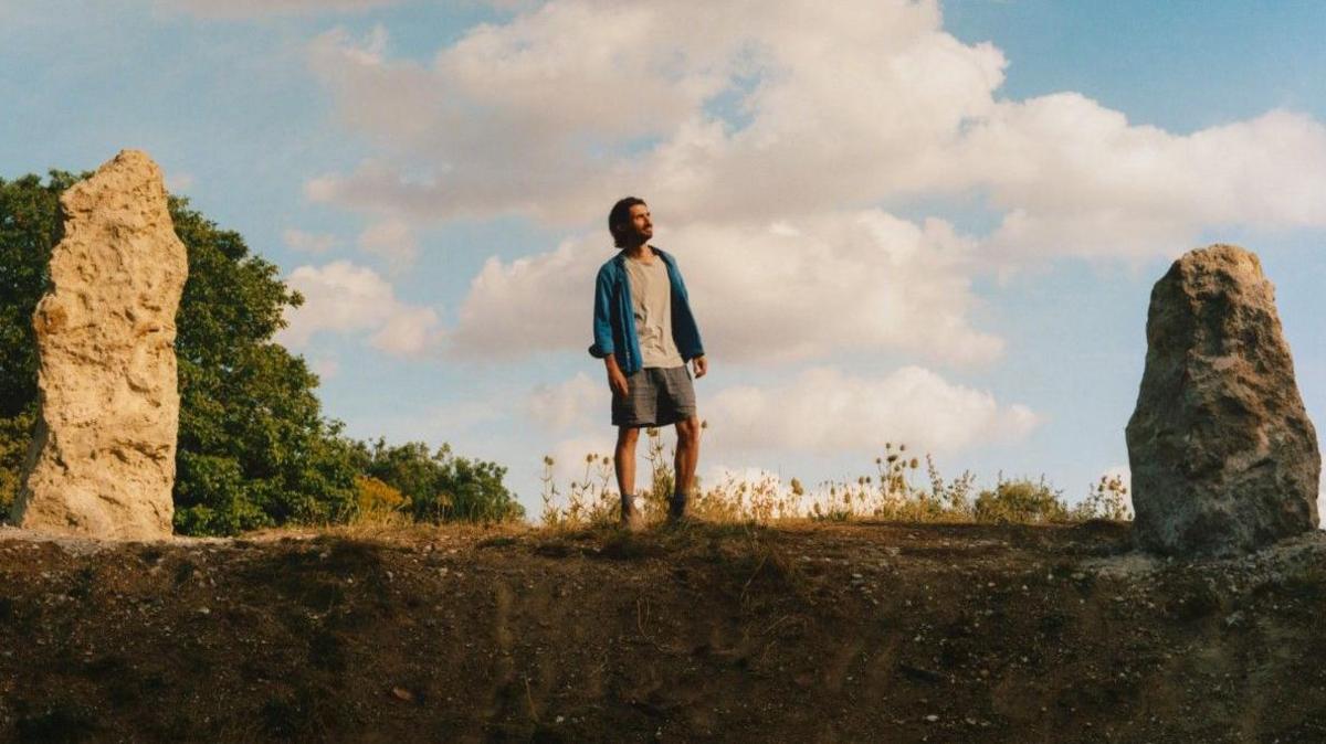 Matthew Rosier is standing on a grass verge in the middle looking towards the right. He is wearing a white top, blue jacket, and grey shorts, and has brown hair. Either side of him are chalk stones and trees in the background.