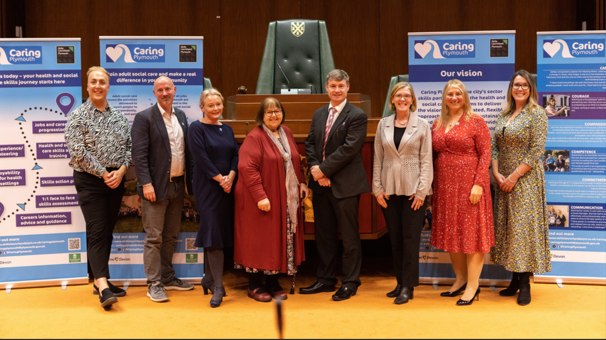 People standing in the Plymouth City Council chamber with Caring Plymouth posters. Left to right the people are Jack Davies, Health and Social Care Coordinator; Neil Eastwood, author of 'Saving Social Care'; Emma Crowther, Interim Head of Commissioning at Plymouth City Council; Councillor Mary Aspinall, Cabinet Member for Health and Adult Social Care; David McAuley, Programme Director of Plymouth Local Care Partnership; Tracey Lee, Chief Executive of Plymouth City Council; Emma Hewitt, Skills Lead at Plymouth City Council, Lauren Parker-Smith, Health and Social Care Coordinator. 