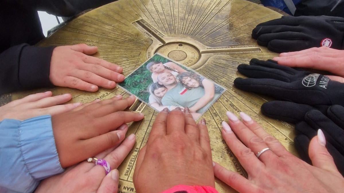 Friends and family of James Cook putting their hands around a photo of him, which is resting on the summit of Snowdon.
