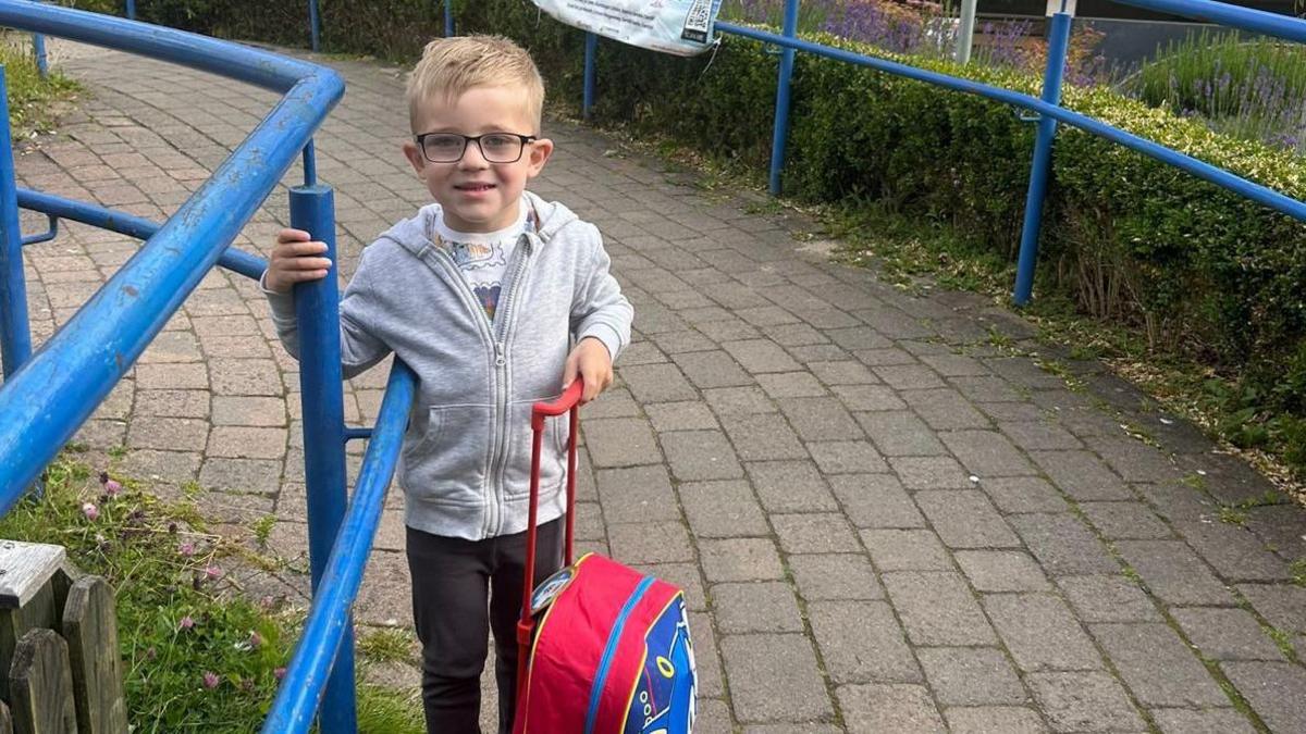 A young boy with blonde hair wearing glasses, a grey jacket and black trouser. He is holding a little red suitcase and smiling at the camera. 