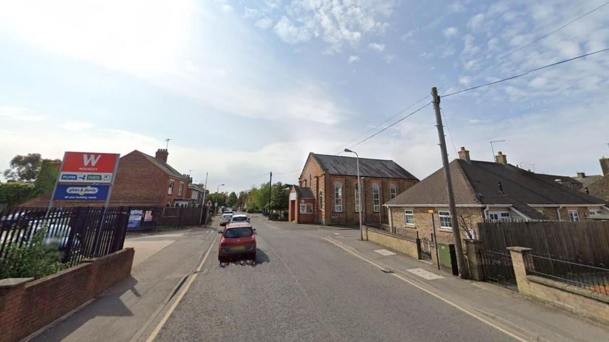 Google street view of a junction with a business on one side with a sign and a car park and a bungalow and church on the other side. A few cars can be seen on the road.