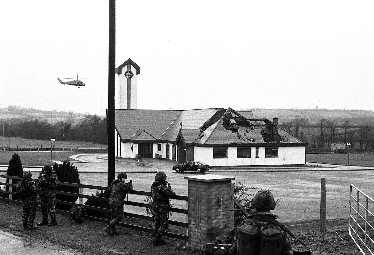 Five soldiers wearing camouflage gear stand guard beside the carpark of St Patrick's Church, which has a partially crumbling rook. A car sits stationary in the car park while a helicopter flies overhead. 