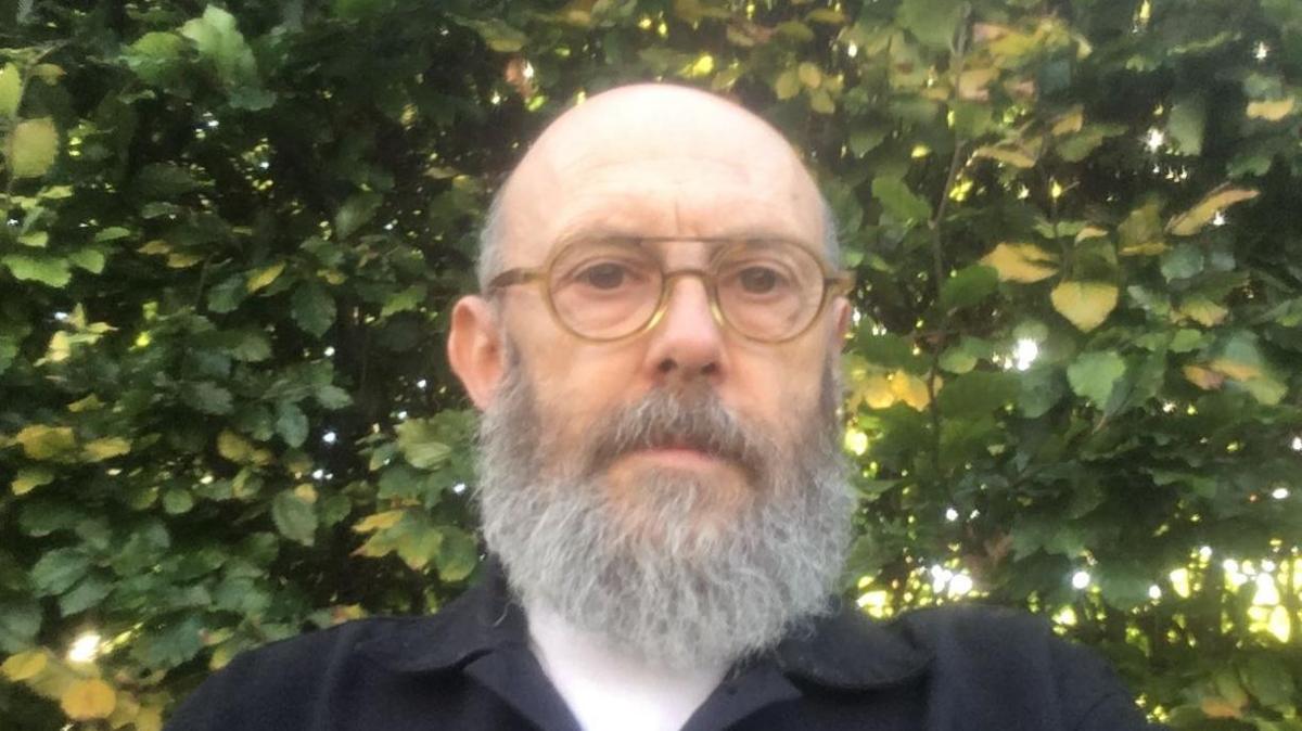 A photo of Andrew Stringer. He is pictured in front of a bush. He is facing the camera with no expression on his face. He is wearing a black coat with a white top underneath. He has a long grey beard, he is bald and is wearing glasses. 