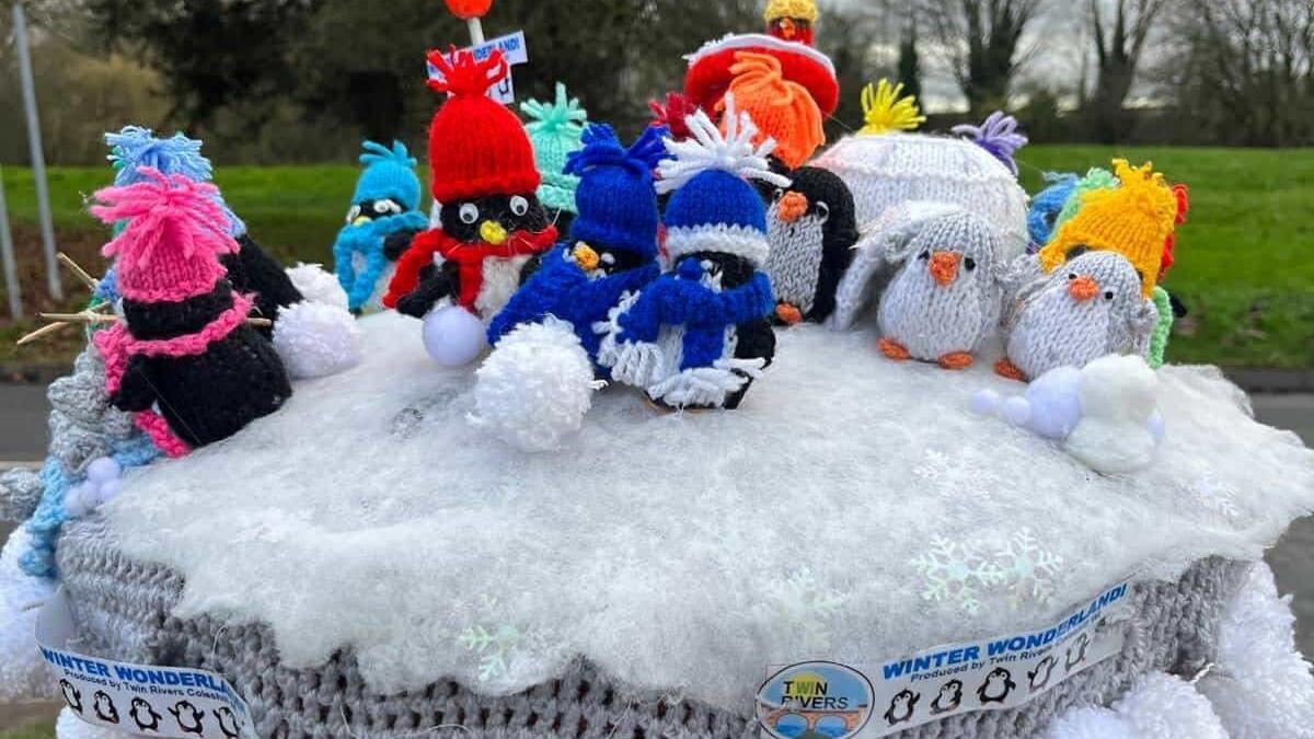 Several skating penguins show them wearing brightly coloured scarves and hats in pink blue and yellow. There are snowball pompoms and baby penguins in grey to one side. They all stand on a fluffy white surface, representing snow.