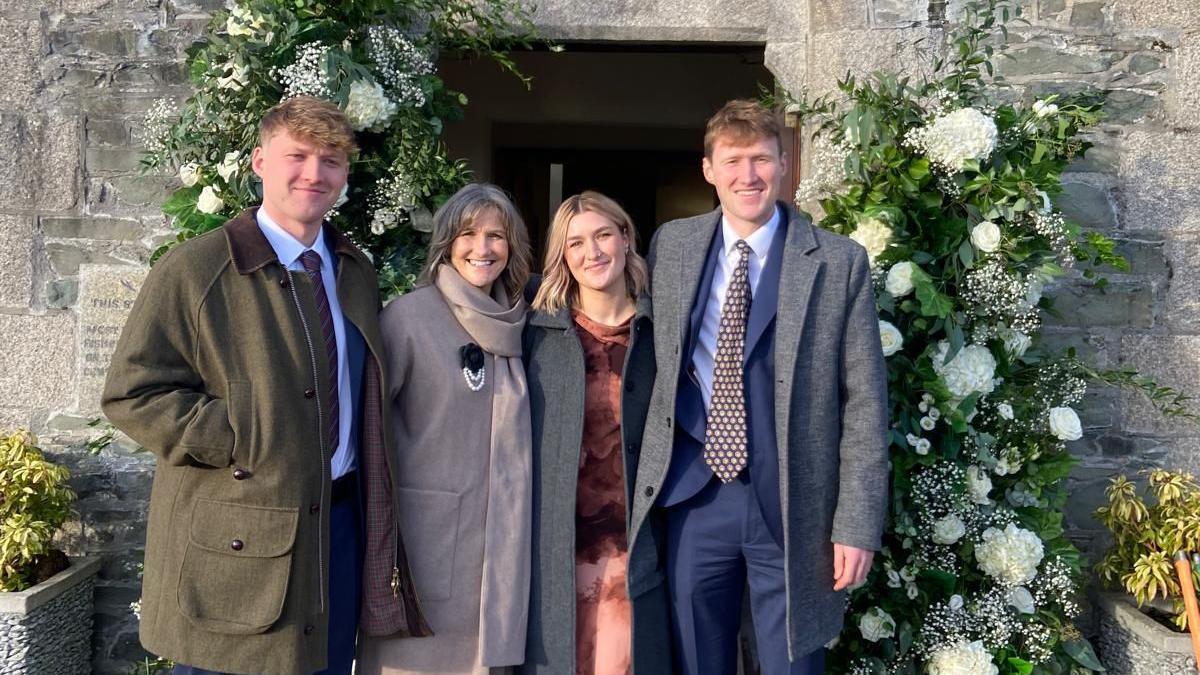 A family of four pose arm-in-arm in front of an open doorway lined with white flowers set in a wall of grey stone. From left to right is a young man with red hair wearing a green jacket above a white shirt and purple tie, a woman with grey hair wearing a beige jacket and scarf, a young woman with blonde hair wearing a patterned orange dress, and a young man with red hair wearing a grey jacket above a blue suit with a patterned brown tie.