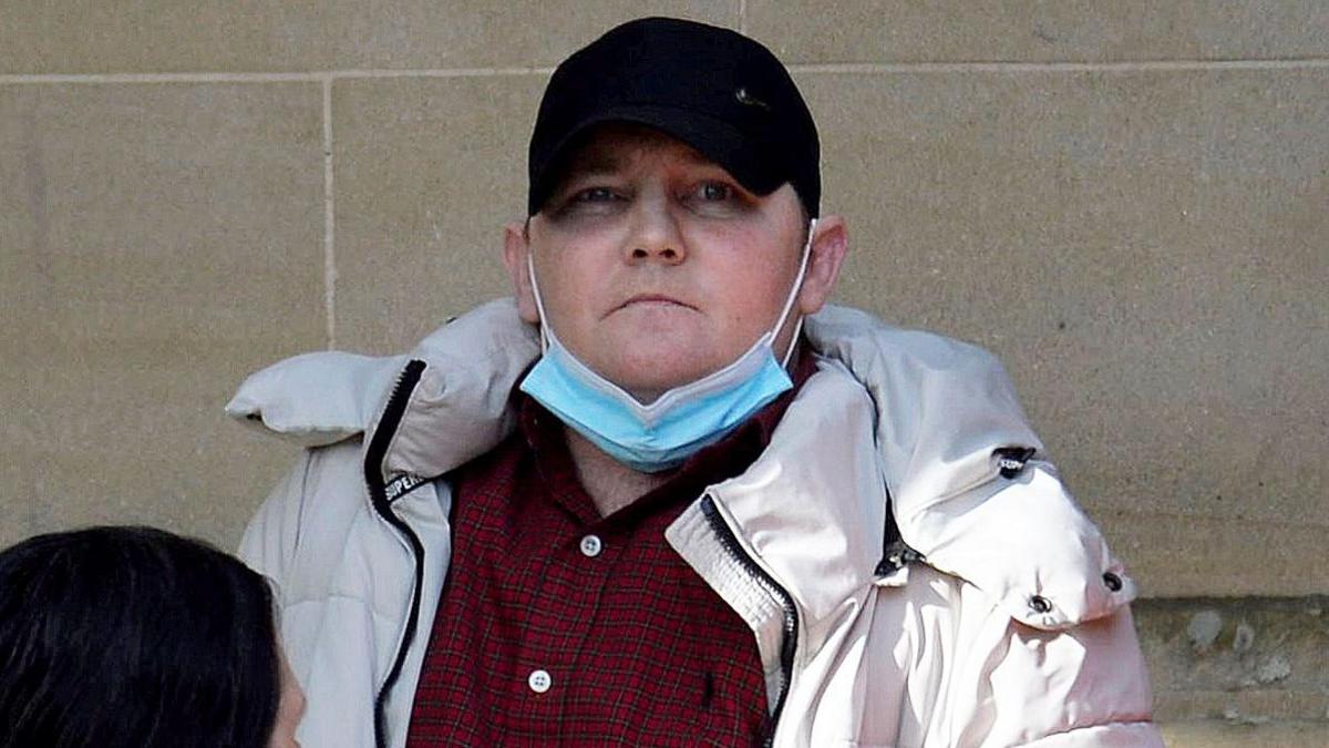 Iain Owens photographed outside the High Court in Glasgow. He is wearing a black baseball cap, red checked shirt, white quilted jacket with a hood. He has a blue and white disposable face mask hanging from his ears and pulled down over his chin. Owens is staring straight at the camera and has a serious expression on his face.