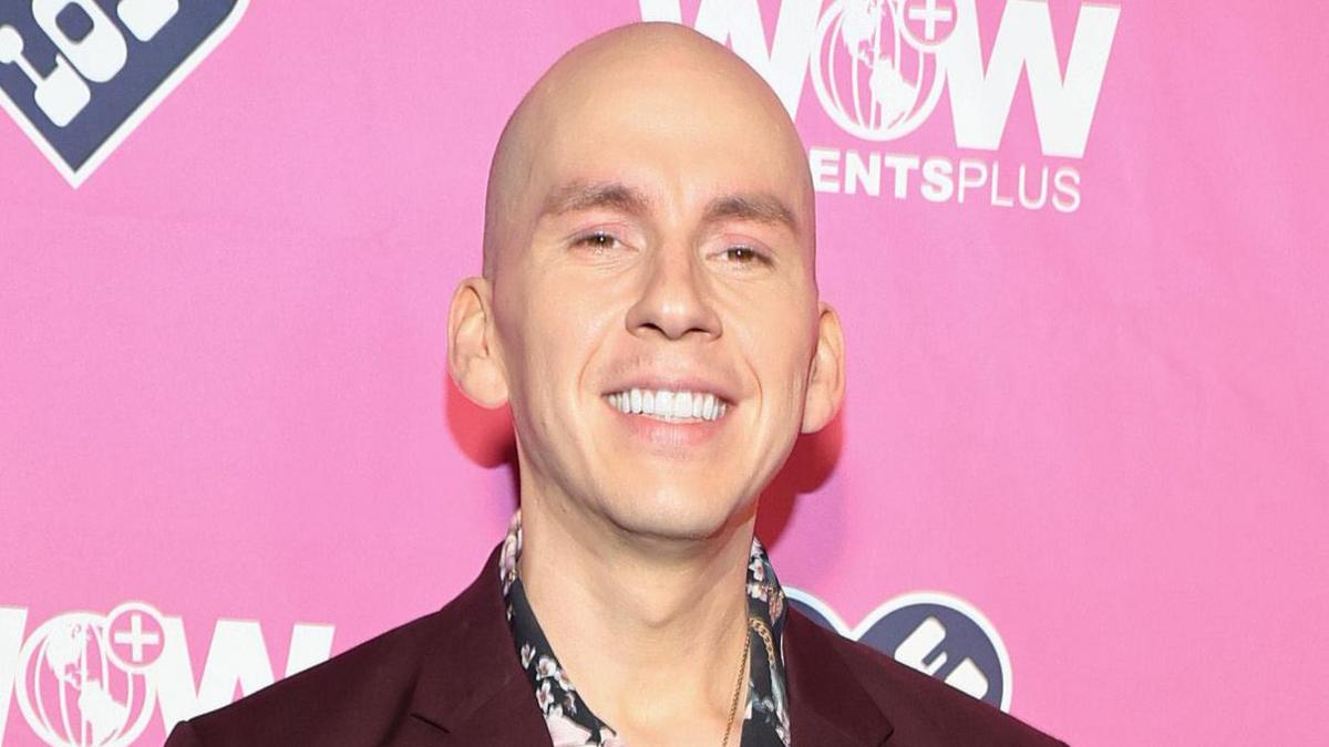 Brian Firkus out of drag. Brian is a bald man in his 30s with brown eyes. He wears a maroon blazer over a patterned black shirt and smiles at the camera 