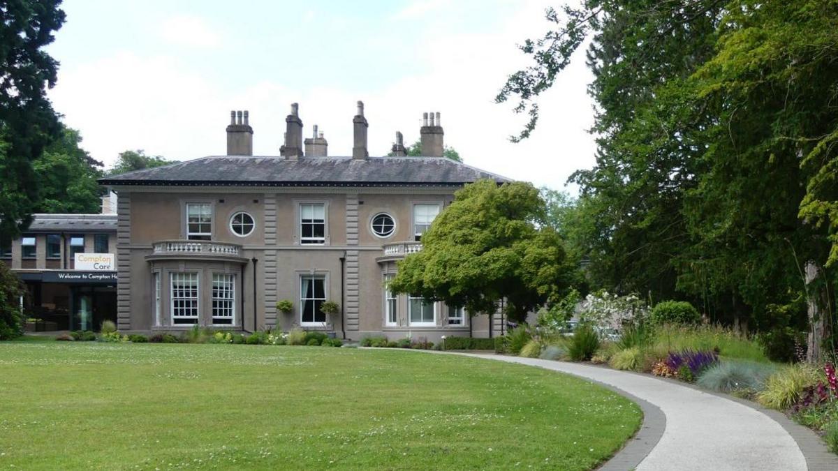 A two-storey brick building, which has large bay windows and looks like a stately home, with a curved path approaching it. A more modern building can be seen behind it on the left.