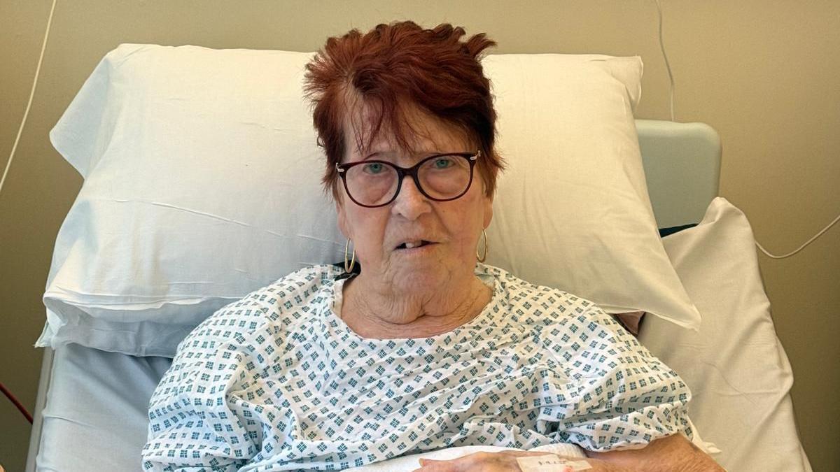 Woman with short red hair and glasses in hospital gown lying in hospital bed