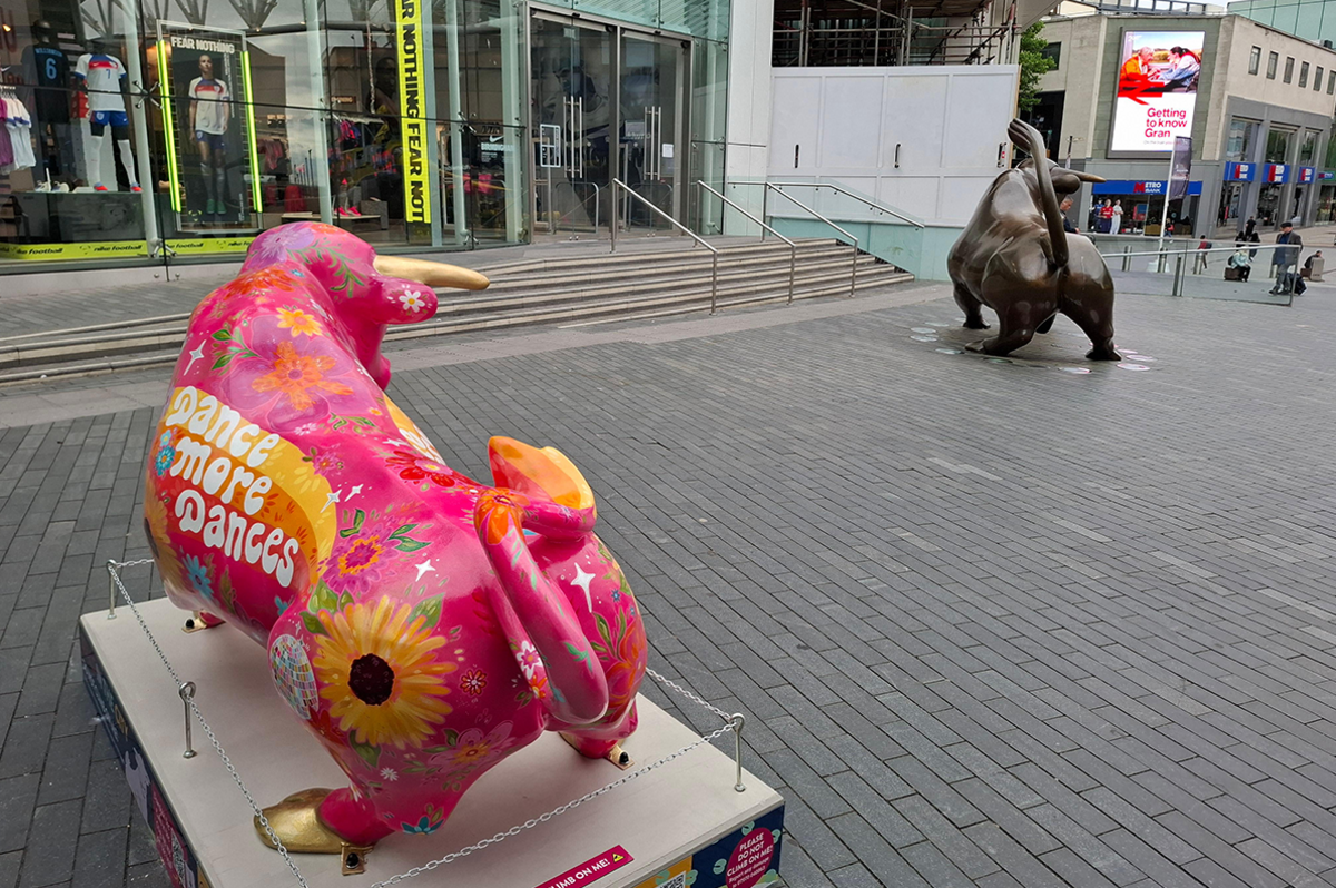 A statue of bull painted pink and featuring yellow flowers on a white plinth on block paving near the Bullring shopping centre. On it is the slogan "Dance more dances". To the right is the shopping centre's resident bronze sculpture 