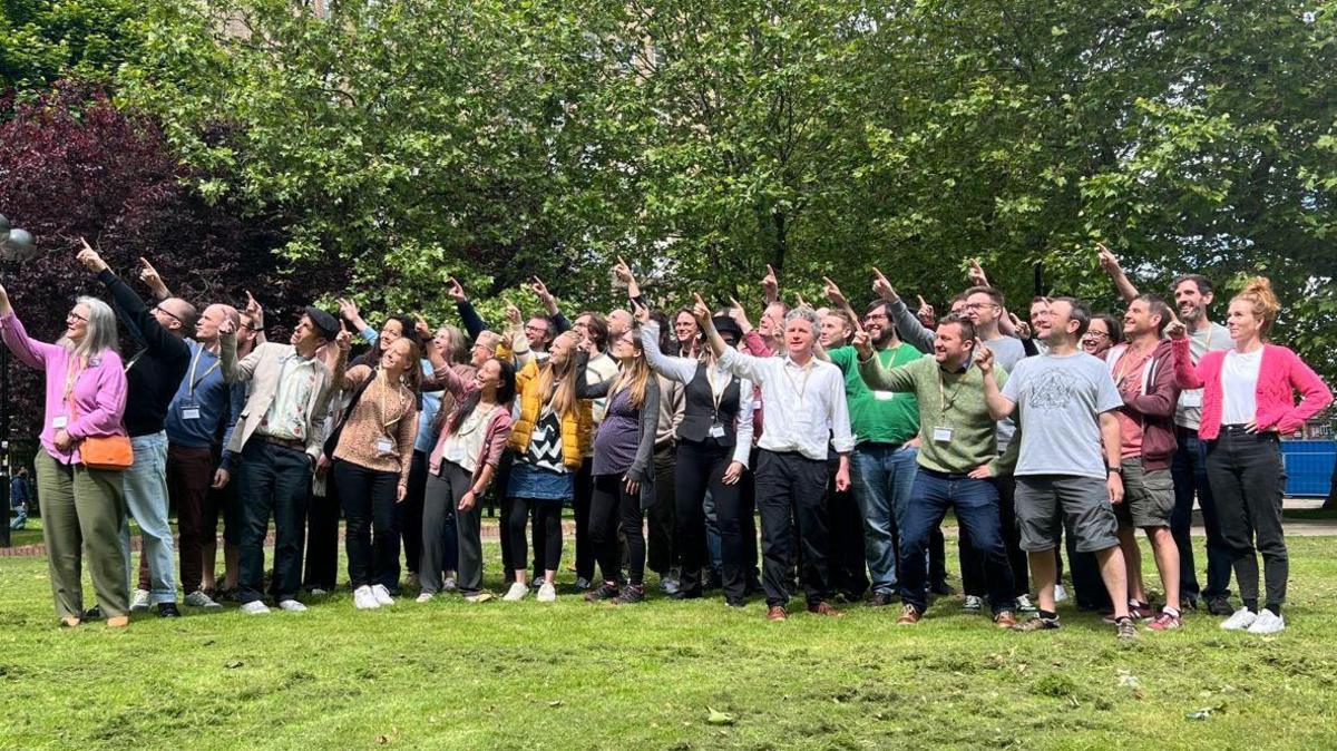 A group photo of Prof Goldacre and the OpenSAFELY research team taken outside on grass with a large tree behind them. They are all pointing right, looking upwards and smiling. Prof Goldacre is pointing up and looking at the camera.