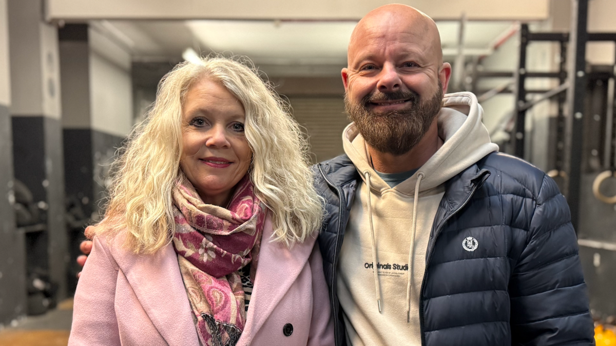 A woman with blonde hair wearing a light coloured scarf and a pink coat stands next to her husband who is wearing a grey coloured hoody underneath a blue jacket.