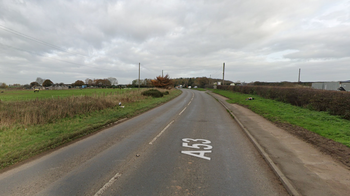 A Google Streetview screengrab of the crash site. A road stretches away into the distance, with A53 superimposed on to it. On the right hand side there's a pavement, and a grassy verge and hedgerow. On the left is a grassy verge with a short hedge with farmland visible beyond it. The sky is grey and overcast. There are bunches of flowers lying either side of the road - the tributes to both men who died. 