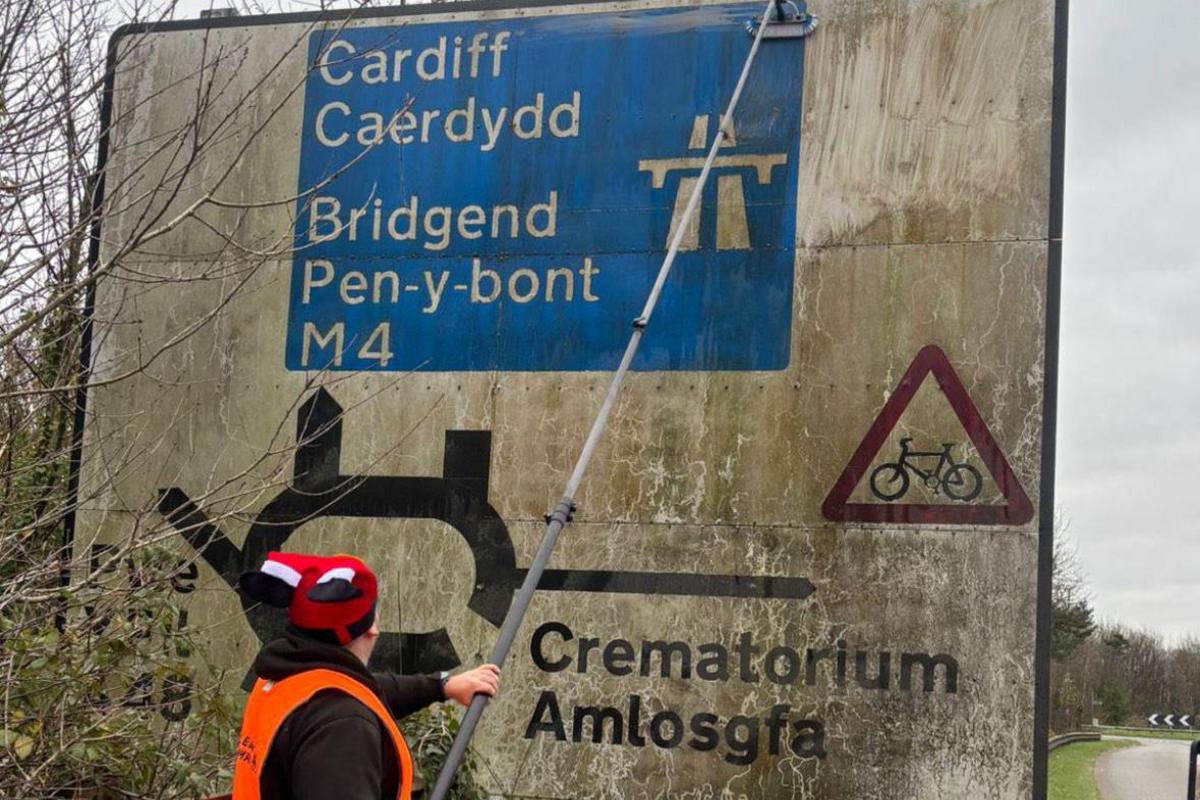 Man in front of a road sign cleaning it with window cleaning equipment. He is stood next to a busy main road.