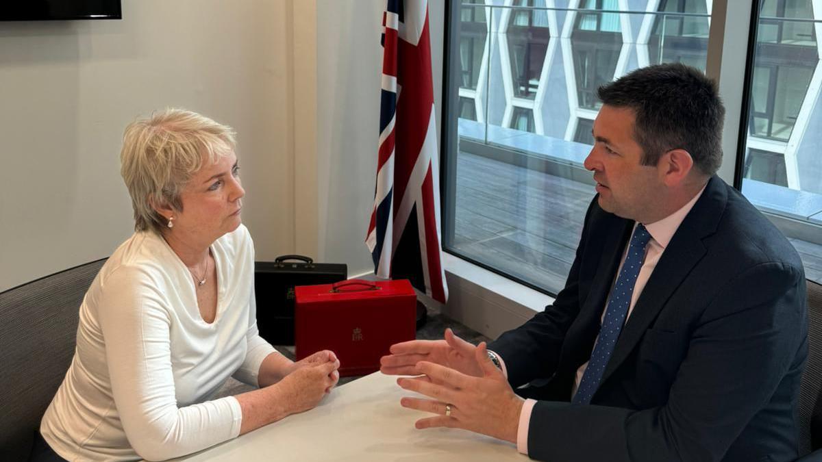 Shaun Davies MP and health minister Karin Smyth talking at a table in Parliament with a union flag on a flag pole behind them, plus brief cases