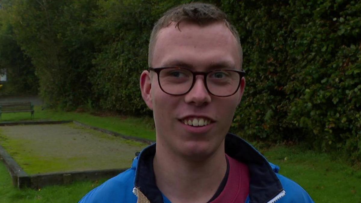 Callum McGinley is wearing a blue jacket, he has short hair, black glasses and is smiling at the camera.