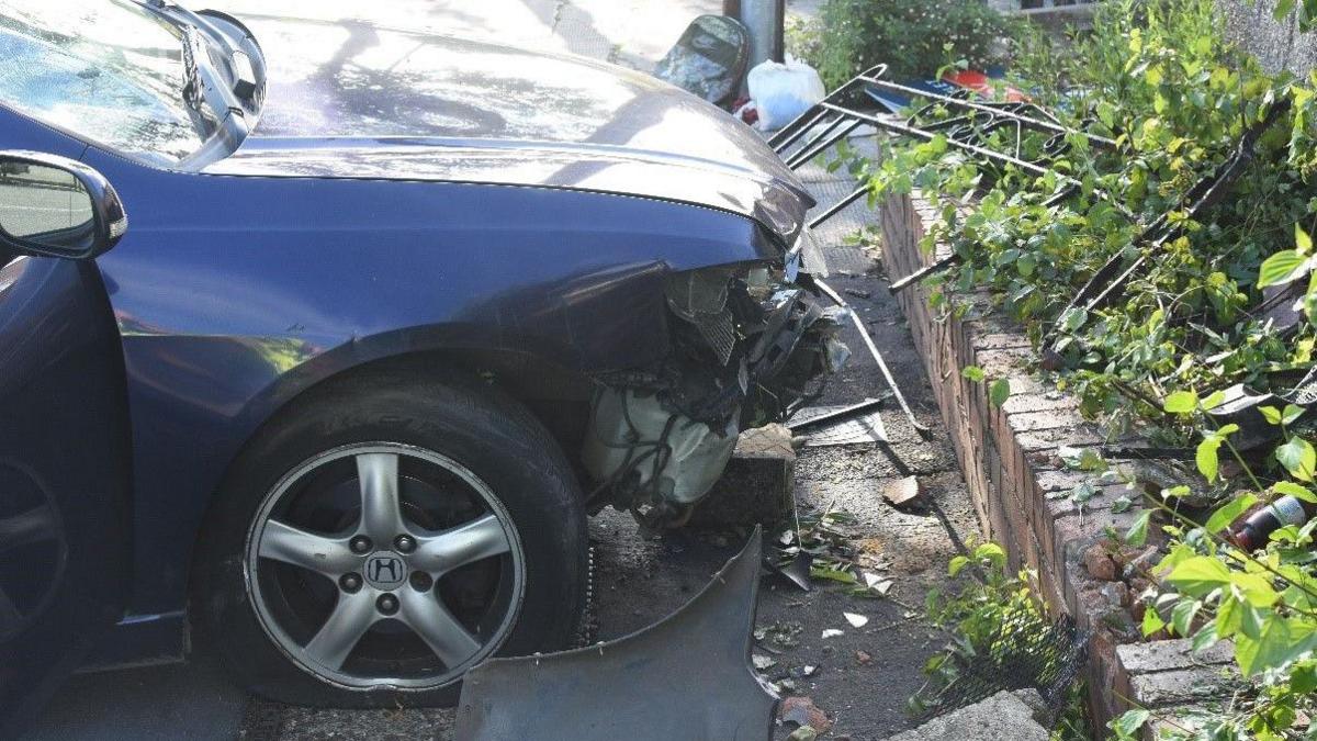 The front of the car which has been mounted onto the pavement. The bumper is smashed and crumpled from the force of the impact. There is also an old bed frame which has been crumpled and is lying next to the low brick wall which Mr Tjitendero was pinned against.