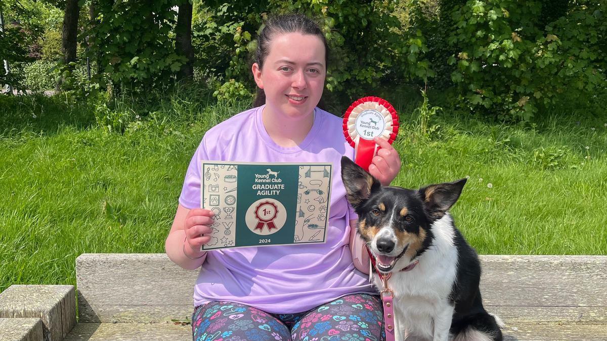 Jessica and her collie dog Zola sit on a park bench. Jessica is wearing a pink t shirt and is holding a certificate and rosette, won by Zola. Zola sits to her left, a pink lead can be seen hanging around her neck