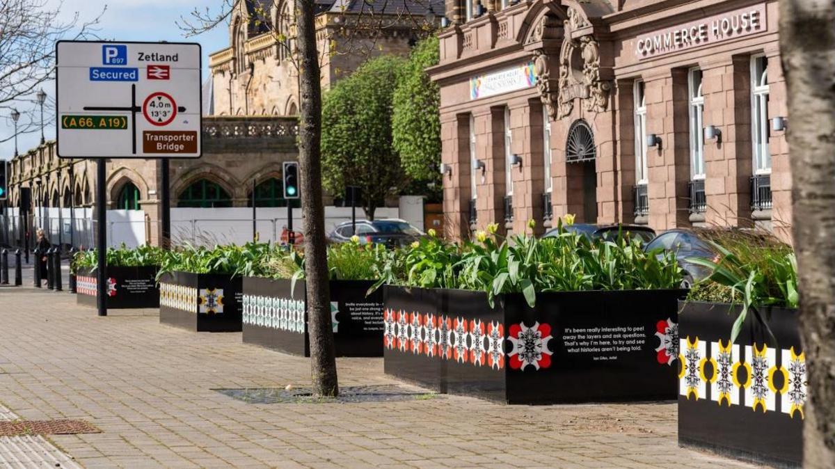 A pedestrianised street, with to the right an imposing Victorian sandstone building fronted by raised modern planters with a patterned design on the front and containing flag irises. The ornate frontage of Middlesbrough Station can be seen to the left, with fencing and white barriers around it.
