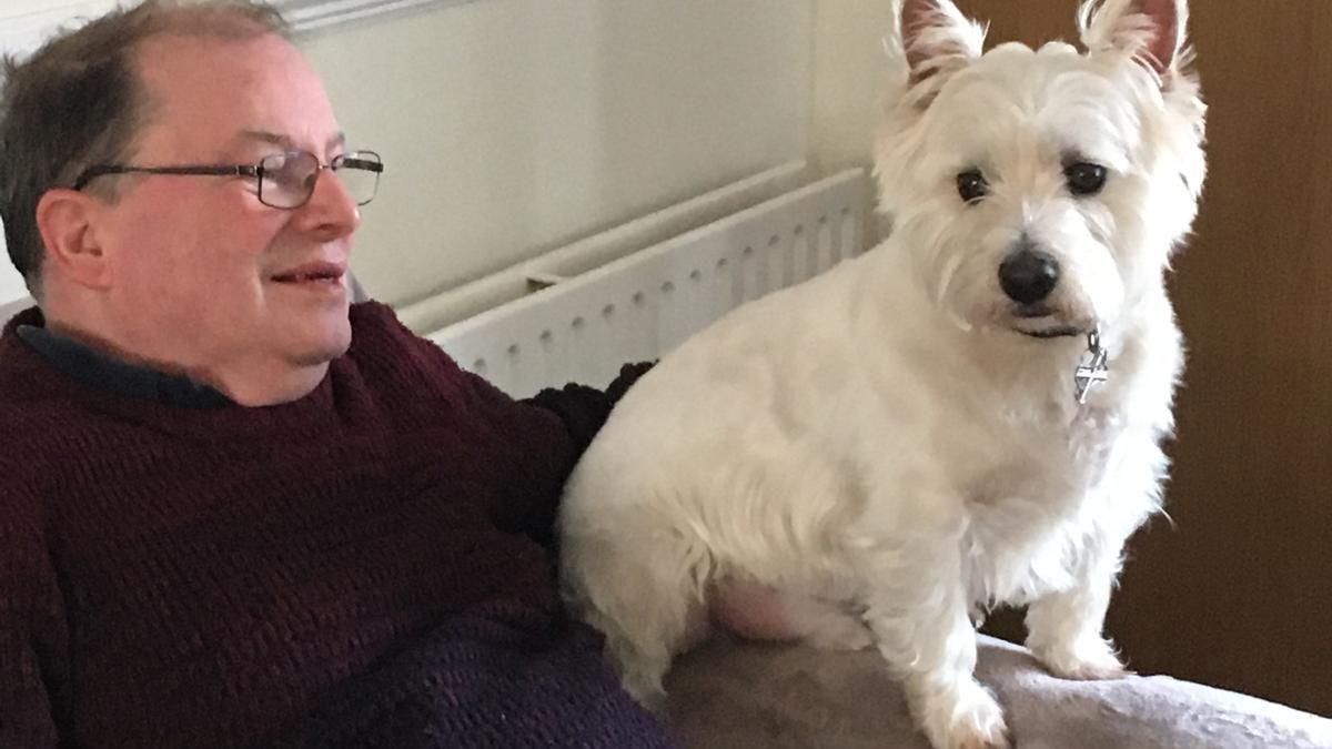 A man sits on a sofa with a short white-furred dog perched on the arm of the sofa. The man wears thin-rimmed, rectangular glasses, has short dark hair and wears a dark red jumper. Behind them is a white radiator.