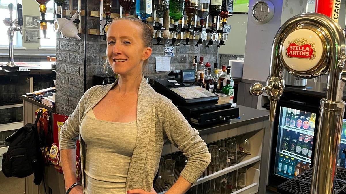 Antonia Merner, with blonde hair tied back in a bun and a grey cardigan over a grey top, smiles at the camera behind the bar.