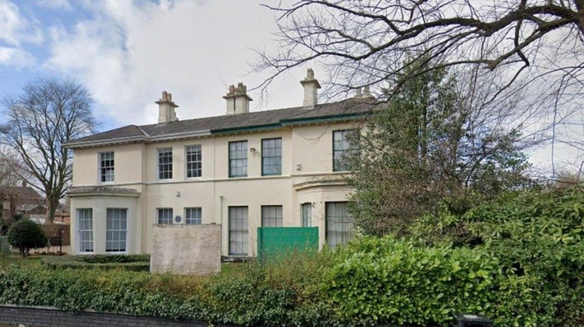 A light-coloured two-storey building has a tree to the left of it on the photo. A hedge is above a small brick wall in the foreground.