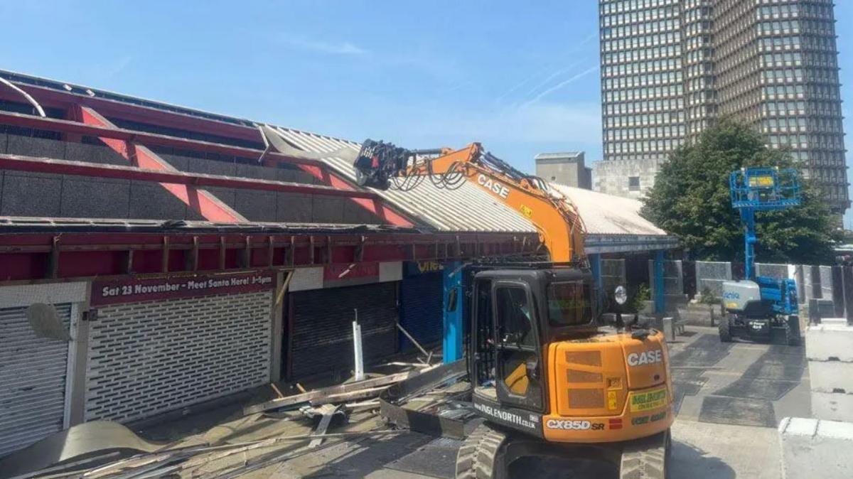 A yellow demolition vehicle removes corrugated roof panels from a single storey structure