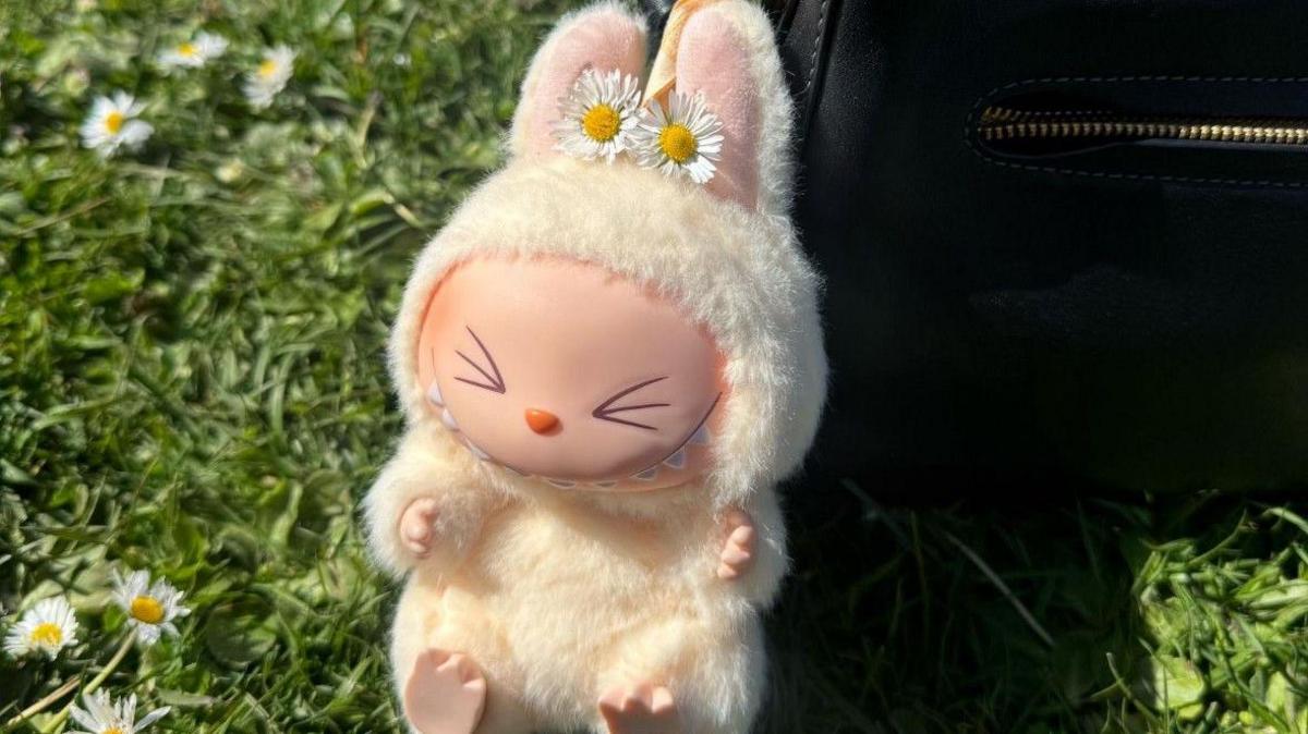 A yellow Labubu bag charm attached to a black bag that is sat on grass, the Labubu has two daisy placed on its ears