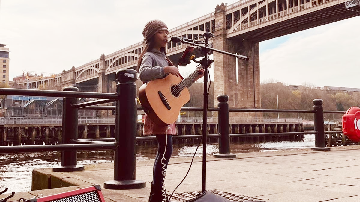 Nine-year-old Skye busking on Newcastle Quayside. She is wearing a beanie hat and is playing an acoustic guitar. She is singing into a microphone on a stand. The River Tyne and the High Level Bridge are in the background behind her.
