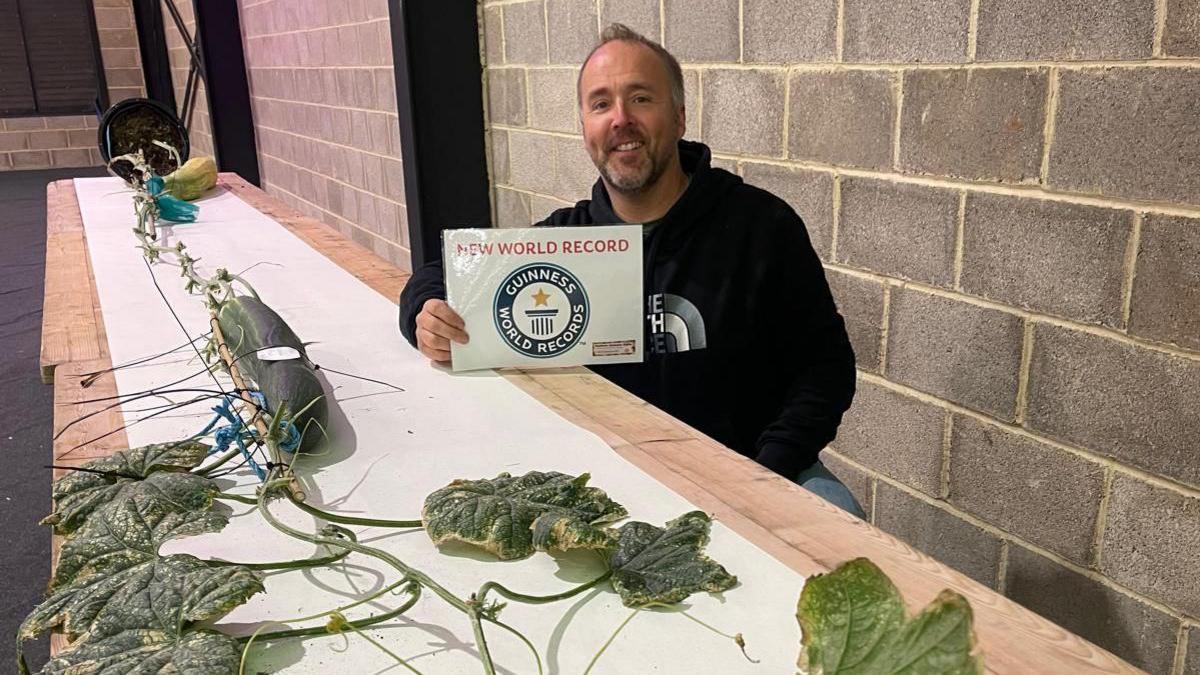 A man holding a Guinness World Record certificate, with plants crawling over a table in the foreground. 