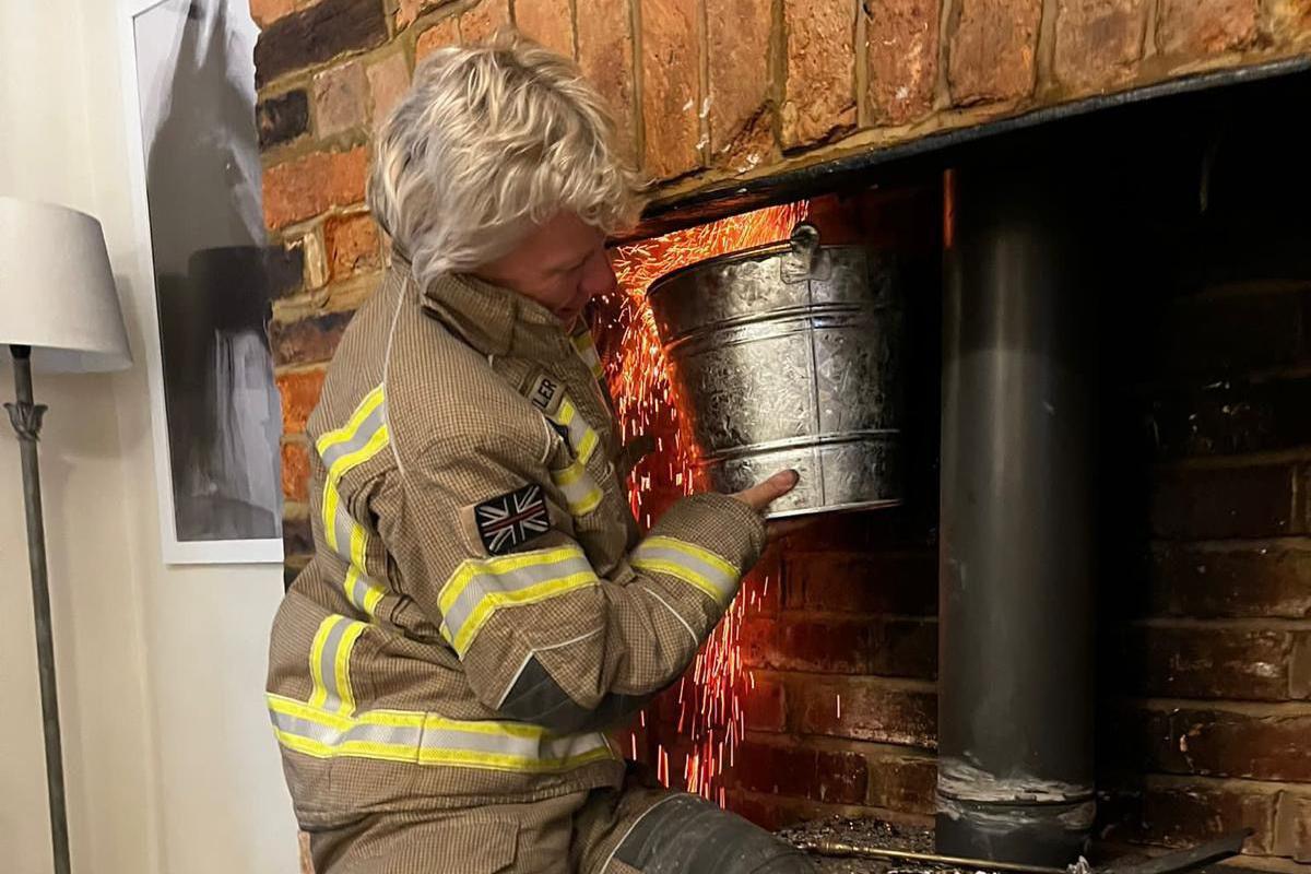 Sally Shayler wearing brown and yellow protective gear stands by a brick fireplace, holding a metal bucket inside the chimney with sparks flying. The floor is covered with a tarp, and firewood is stacked nearby. This shows chimney maintenance with safety precautions.