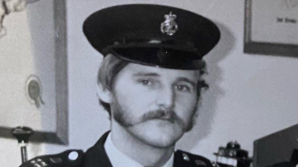 A black and white image of a male police officer in uniform. He has a thick, wide moustache leading into sideburns. He is wearing a black peaked police cap with a badge in the centre. A white collar and black jacket can be seen, with  shoulder epaulettes visible, He is standing in front of a white wall with undiscernible office equipment behind.