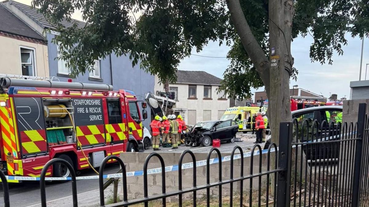 A Staffordshire Fire Service vehicle at the scene of a crash. A damaged black car can be seen in the distance, and an ambulance. Emergency service staff are at the scene, and a collapsed building can be seen behind them.