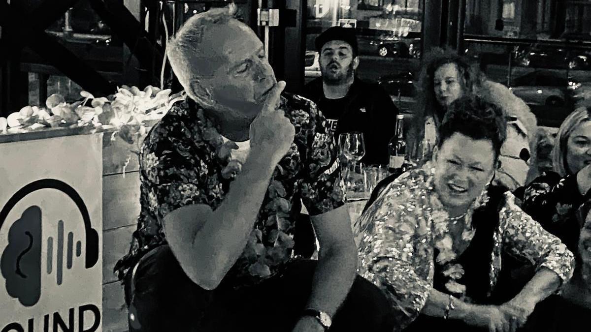 Fatboy Slim, AKA Norman Cook, wearing a floral short-sleeve shirt, sits with group of people discussing DJ mixing at the Sounds Mind event. It is a black and white photograph.